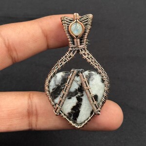 Puede incluir: Un colgante artesanal con una piedra en forma de corazón con patrones en blanco y negro, engarzada en alambre de cobre. El colgante incluye una pequeña piedra redonda e iridiscente en la parte superior. El trabajo del alambre es intrincado, creando un diseño artístico único.