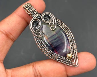 May include: A handcrafted pendant featuring a polished, oval-shaped gemstone with purple, green, and white hues. The stone is encased in a woven silver-colored wire frame with decorative swirls and a small gold-colored bead.