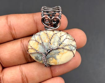 May include: Oval pendant featuring a wire-wrapped tree design. The pendant has a light-colored stone base with a detailed copper wire tree and bail. The stone has yellow, gray, and white tones. The bail is made of dark wire.