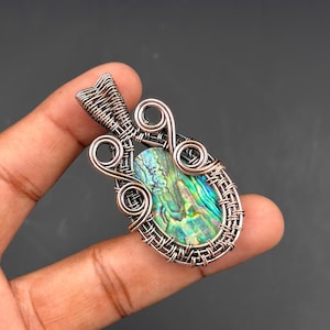 Op de afbeelding: Ovale hanger met een levendig abalone schelp centrum, omhuld door ingewikkelde draadwikkeling. Het draadwerk heeft zilver- en kopertinten, wat een uniek en artistiek ontwerp creëert. De hanger wordt in een hand gehouden tegen een donkere achtergrond.