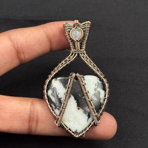 Puede incluir: Colgante artesanal con una piedra en forma de corazón con patrones en blanco y negro, engarzada en alambre de cobre. El trabajo de alambre incluye una pequeña piedra redonda en la parte superior. El colgante es sostenido por una mano sobre un fondo oscuro.