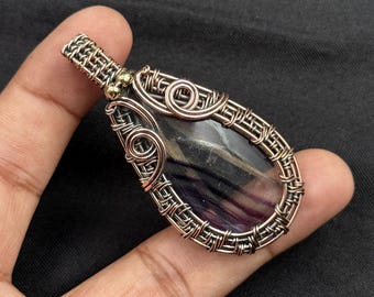 May include: A handcrafted pendant featuring a teardrop-shaped stone with purple and gray hues, encased in intricate copper wire. The wirework includes decorative swirls and a bail for a necklace chain.
