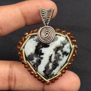 Puede incluir: Colgante en forma de corazón con una piedra central en blanco y negro. La piedra está enmarcada con alambre dorado y pequeñas cuentas marrones. Una espiral y una fianza de alambre plateado completan el colgante.