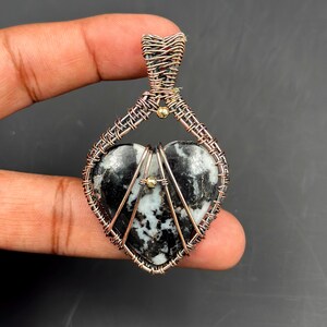 Puede incluir: Colgante en forma de corazón con una piedra central en blanco y negro, envuelto intrincadamente en alambre color cobre. El alambrado forma una jaula alrededor de la piedra y un lazo para una cadena. Pequeñas cuentas doradas acentúan el diseño. Una joya única.