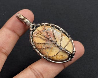 May include: Oval pendant with a beige and brown stone, wrapped in copper wire. The wire is shaped to resemble a tree. The bail is silver and gold-toned wire. The pendant is held in a hand.