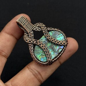 Op de afbeelding: Een handgemaakte hanger met een abalone schelp, ingekapseld in ingeweven koperdraad. De hanger toont iriserende blauwe, groene en paarse kleuren in de schelp, wat een uniek sieraad creëert.
