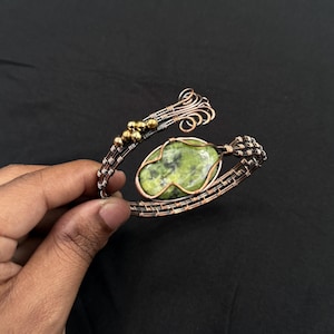 Peut inclure: Un bracelet artisanal en fil de cuivre avec une grosse pierre verte ovale. Le bracelet a un design tissé avec des perles dorées et des volutes décoratives. Le bracelet est présenté sur un fond sombre.
