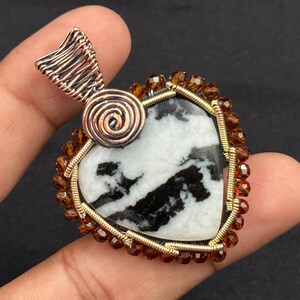 Puede incluir: Colgante artesanal con una piedra en forma de corazón con patrones en blanco y negro. La piedra está enmarcada con alambre dorado y adornada con pequeñas cuentas marrones. El colgante tiene una fianza de alambre de cobre y un acento en espiral.