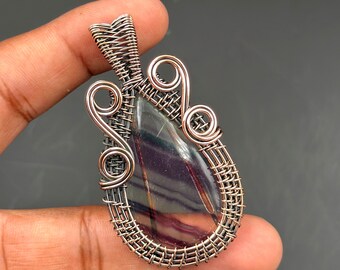 May include: A handcrafted pendant featuring a teardrop-shaped, multi-colored stone, encased in a woven silver-toned wire frame. The pendant has decorative swirls and a bail for a chain. The stone displays shades of purple, green, and brown.