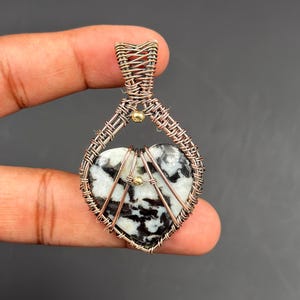 Puede incluir: Colgante en forma de corazón con una piedra negra y blanca en el centro, envuelto en alambre de color cobre. El trabajo de alambre forma una jaula alrededor de la piedra, con un lazo para una cadena. Pequeñas cuentas doradas acentúan el diseño.