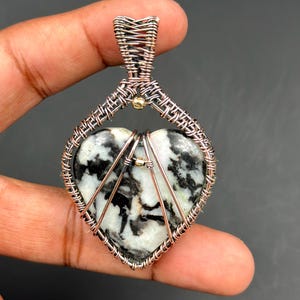 Puede incluir: Un colgante en forma de corazón con un centro de piedra en blanco y negro, intrincadamente envuelto en alambre de cobre. El colgante está suspendido de una fianza de alambre tejido y presenta pequeños acentos dorados. El diseño se sostiene en una mano.