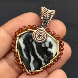 Puede incluir: Colgante en forma de corazón con una piedra central en blanco y negro. La piedra está enmarcada con alambre dorado, pequeñas cuentas naranjas y una fianza de alambre de cobre. Un detalle en espiral de cobre se encuentra en la parte superior de la piedra.