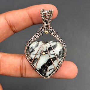 Puede incluir: Un colgante en forma de corazón con una piedra en blanco y negro, encerrado en un marco de alambre color cobre. El alambre está intrincadamente tejido, con detalles dorados. El colgante se sostiene en una mano sobre un fondo oscuro.