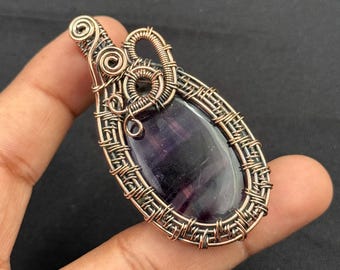 May include: A handcrafted pendant featuring a polished, oval-shaped purple gemstone, intricately wrapped in copper wire. The wirework includes decorative swirls and a woven pattern, creating a unique, artistic design. The pendant is designed to be worn as a necklace.