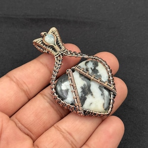 Puede incluir: Colgante artesanal con una piedra en forma de corazón con patrones en blanco y negro, envuelta en alambre de cobre. El colgante tiene una pequeña piedra azul claro en la parte superior. El trabajo del alambre es intrincado y detallado, creando una joya única.
