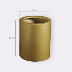 May include: A cylindrical gold-colored planter, 30 cm tall and 25 cm in diameter. The planter has a smooth, glossy finish and a wide opening. The logo "JD JOIA DECO & STYL" is visible on the front.