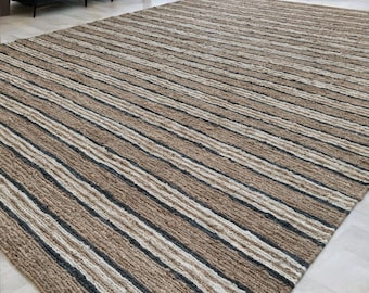 Può includere: Un grande tappeto rettangolare con un motivo a righe nei toni del marrone, beige e grigio scuro. Il tappeto è realizzato in un materiale tessuto e si trova su un pavimento in legno chiaro. Il tappeto è un elemento di arredo per la casa.