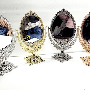May include: Four oval tabletop cosmetic mirrors with ornate, decorative frames and rectangular bases. The mirrors are finished in silver, gold, dark silver, and rose gold metallic tones. Each mirror is mounted on a swivel stand for adjustment.
