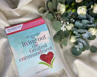 May include: A book titled "Life principles for living out the Greatest commandment" by Cheri Strange. The book has a red heart graphic and is next to a bouquet of white roses and greenery on a cream-colored fabric.