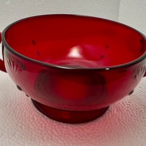 Könnte beinhalten: Eine rote Glasschüssel mit zwei Griffen. Die Schüssel hat eine strukturierte Oberfläche und einen kleinen Sockel.