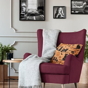 Puede incluir: Un sill&oacute;n burdeos con una manta de lana gris y un coj&iacute;n decorativo con un patr&oacute;n de Halloween de gatos negros y calabazas naranjas.