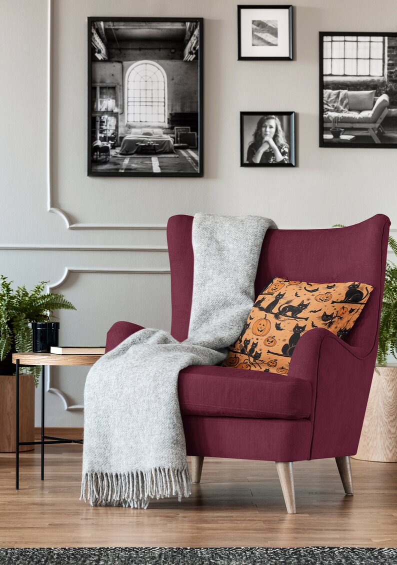 Puede incluir: Un sill&oacute;n burdeos con una manta de lana gris y un coj&iacute;n decorativo con un patr&oacute;n de Halloween de gatos negros y calabazas naranjas.