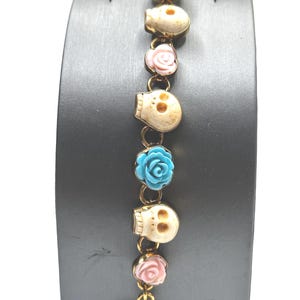 May include: Gold-toned bracelet featuring a repeating pattern of cream-colored skulls and colorful floral accents. The bracelet includes light blue and pink roses, connected by gold-colored links. The clasp is a simple bar and loop design.