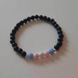 May include: A beaded bracelet featuring black, round beads with a section of light blue, pink, and white beads. The bracelet is strung on an elastic cord and is displayed on a white surface.