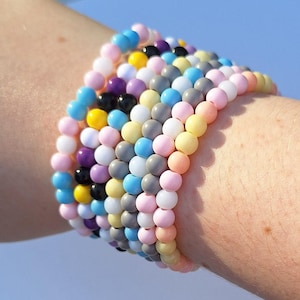 Puede incluir: Una pila de pulseras de cuentas de colores en tonos rosa, azul, amarillo, morado, negro, blanco y gris. Las pulseras se llevan en una muñeca sobre un fondo azul claro. Las cuentas son pequeñas y redondas.