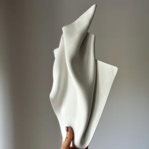 May include: A white, abstract sculpture with a flowing, draped design. The sculpture has sharp, angular points and soft, curved folds, creating a dynamic and textured appearance. The piece is held by a hand, showcasing its form against a neutral background.