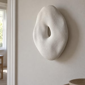 May include: A wall-mounted sculpture featuring an irregular, organic, pebble-like shape with a central aperture. The object has a matte, off-white, textured surface that appears porous and stone-like, creating a three-dimensional form.