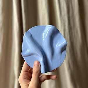 Op de afbeelding: Een lichtblauwe, keramische, decoratieve schaal met een gegolfd oppervlak. De schaal is rond en heeft een glanzende afwerking. De schaal wordt door een hand vastgehouden tegen een neutrale achtergrond. Ongeveer 15 cm in diameter.