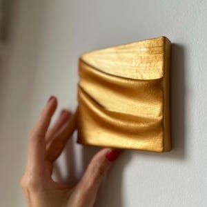 Peut inclure: Une sculpture murale de couleur dorée avec un design ondulé et abstrait. La sculpture est montée sur un mur blanc.