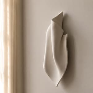 Könnte beinhalten: Eine vertikale Wandskulptur aus cremeweißem Keramikmaterial mit einer gefalteten, stoffähnlichen Optik, glatten, geschwungenen Kanten und einer matten Oberfläche. Das Objekt ist flach an einer Wand in neutralem Farbton montiert.