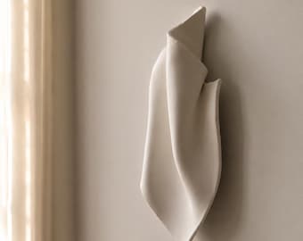 May include: A wall-mounted off-white ceramic sculpture featuring a folded, fabric-like appearance with smooth, flowing curves and a matte finish. The object has a vertical orientation with a pointed bottom and a layered, overlapping structure.