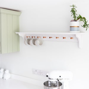 May include: White kitchen shelf with wooden pegs, holding white mugs. A potted succulent plant sits on the shelf. A white stand mixer is on the counter below.