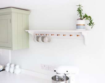 May include: White kitchen shelf with wooden pegs, holding white mugs. A potted succulent plant sits on the shelf. A white stand mixer is on the counter below.