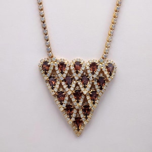May include: A gold-coloured necklace featuring a triangular pendant. The pendant is decorated with teardrop-shaped, dark red gemstones and smaller, white iridescent stones. The necklace chain is also embellished with small, iridescent stones.