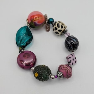 May include: A colorful beaded bracelet with a variety of shapes and textures. The bracelet features beads in shades of pink, teal, black, and green. Some beads have patterns, while others are smooth and glossy. The bracelet is a unique piece of jewelry.