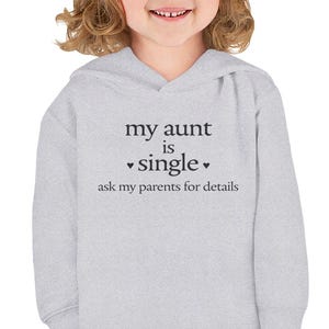 Puede incluir: Una sudadera con capucha de color gris claro y manga larga que presenta el texto en negro "my aunt is single" seguido de dos pequeños iconos de corazón y el texto "ask my parents for details" impreso en el centro.