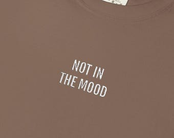 May include: A folded brown t-shirt with the phrase "NOT IN THE MOOD" printed in white. The shirt has a crew neck and a tag on the inside collar.