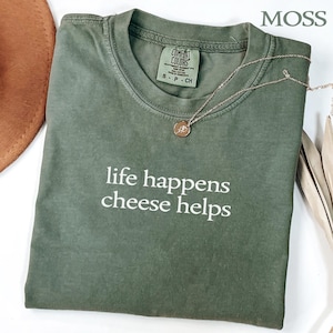 Könnte beinhalten: Ein moosgrünes T-Shirt mit Rundhalsausschnitt und dem weißen Schriftzug life happens cheese helps auf der Vorderseite. Das Shirt hat ein Comfort Colors Markenetikett am Innenkragen mit der Größenangabe small und eine goldene Halskette.