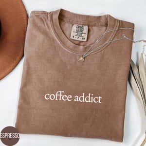 Könnte beinhalten: Espressofarbenes T-Shirt mit dem weißen Schriftzug „coffee addict“. Das Shirt hat einen Rundhalsausschnitt und kurze Ärmel. Eine silberne Halskette mit Anhänger ist über das Shirt drapiert. Das Shirt ist auf einer weißen Oberfläche gefaltet.