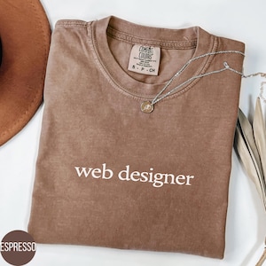 Può includere: Una t-shirt a maniche corte color espresso con la scritta "web designer" stampata in bianco sul petto. La maglietta presenta un'etichetta del marchio Comfort Colors all'interno del colletto ed è abbinata a una delicata collana a catena argentata.