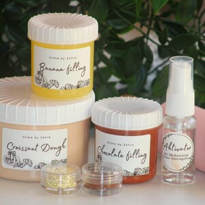 Puede incluir: Una colección de slimes en varios recipientes, incluyendo un recipiente grande con la etiqueta "Croissant Dough" y frascos más pequeños con "Banana filling" y "Chocolate filling". También hay un frasco de spray etiquetado "Aktivator" y un rollo rosa.