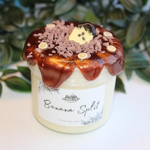 Könnte beinhalten: Ein transparentes Glasgefäß gefüllt mit einer weißen cremigen Substanz, die mit einer braunen Schokoladensauce und Schokoladenstreuseln bedeckt ist. Das Gefäß hat ein weißes Etikett mit dem aufgedruckten Text "Banana Split".
