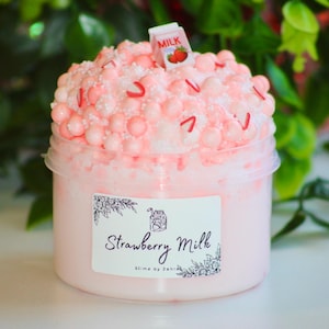 Könnte beinhalten: Ein durchsichtiger Plastikbehälter gefüllt mit rosa Schleim, bedeckt mit rosa Kugeln, roten Streuseln und einem Miniatur-Milchkarton. Das Etikett trägt die Aufschrift "Strawberry Milk" mit floralen Akzenten.