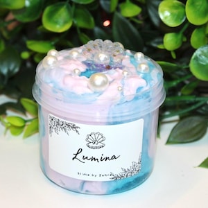 Könnte beinhalten: Ein kleines, durchsichtiges Plastikglas gefüllt mit pastellfarbenem Schleim. Der Schleim ist mit blauen, rosa und lila Farbtönen verwirbelt und mit Kunstperlen und einer dekorativen Muschel verziert. Das Etikett lautet "Lumina Slime by Zahra."