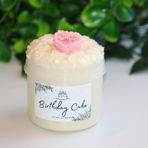 Könnte beinhalten: Ein kleiner, klarer Plastikbehälter gefüllt mit cremefarbenem Schleim, bedeckt mit einer strukturierten, flauschigen Schicht und einer rosa herzförmigen Dekoration. Das Etikett trägt die Aufschrift "Birthday Cake" mit einer Kuchenillustration.