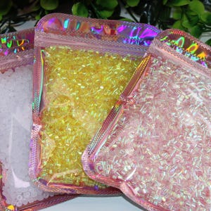 Könnte beinhalten: Drei rosa schillernde Beutel gefüllt mit kleinen Perlen in Weiß, Gelb und Rosa. Die Beutel haben einen holografischen Effekt und sind mit einem Reißverschluss verschlossen. Der Hintergrund ist mit grünem Laub verschwommen.